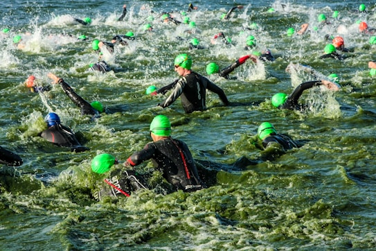 A large group of swimmers wearing green swim caps and wetsuits navigates through turbulent water, creating splashes as they make their way in a competitive swimming event.