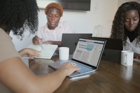 Several people are sitting around a wooden table engaged in a collaborative work session. Laptops and notebooks are open in front of them, with one person writing in a notebook. The screen of one laptop displays a document titled 'modern architecture.' Two white mugs are visible on the table.