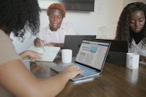 Several people are sitting around a wooden table engaged in a collaborative work session. Laptops and notebooks are open in front of them, with one person writing in a notebook. The screen of one laptop displays a document titled 'modern architecture.' Two white mugs are visible on the table.