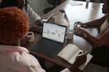 Three people are sitting around a wooden table, each engaged with a laptop. One person is holding a cup of coffee while taking notes in a notebook. The screen of one laptop displays a calendar interface. The setting appears to be a collaborative work environment.