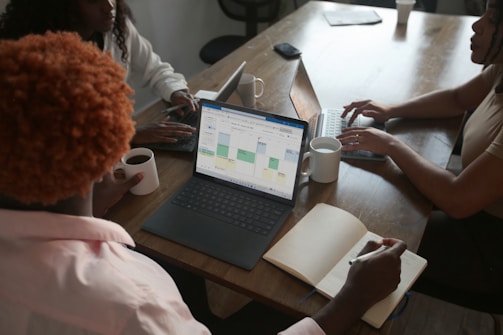 Three people are sitting around a wooden table, each engaged with a laptop. One person is holding a cup of coffee while taking notes in a notebook. The screen of one laptop displays a calendar interface. The setting appears to be a collaborative work environment.