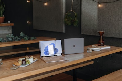 A cozy workspace with a wooden table features two laptops, one open displaying a blue-themed desktop and the other closed. On the table are a tray of snacks, a glass of water, stacked plates, and a decorative coffee maker. The backdrop includes soft wall panels, fairy lights, and plants, creating a warm and inviting atmosphere.