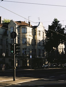 A city street scene with an old residential building illuminated by bright sunlight. The building has multiple windows and decorative architectural features. In the foreground, a traffic light with a green arrow is visible, along with several street signs and power lines crisscrossing overhead. A few cars are parked along the street, and the shadows of trees create patterns on the building facade.