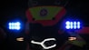 The image displays the front of a vehicle or bike adorned with bright LED lights. Blue and orange lights are prominent next to a central logo or emblem which features a red star and text. The lighting creates a high-contrast effect against the dark background.