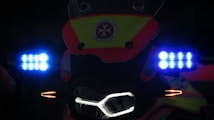 The image displays the front of a vehicle or bike adorned with bright LED lights. Blue and orange lights are prominent next to a central logo or emblem which features a red star and text. The lighting creates a high-contrast effect against the dark background.