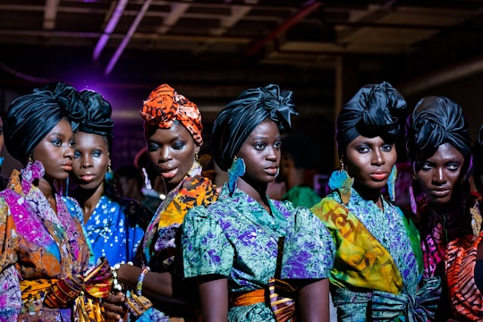 A group of women stands closely together, wearing vibrant, colorful African dresses and headwraps. The outfits feature a variety of patterns, including floral, abstract, and geometric designs. The women have confident expressions and their makeup is elegantly done, accentuating their features. The setting appears to be indoors, possibly under artificial lighting which casts a rich, dramatic effect.