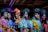 A group of women stands closely together, wearing vibrant, colorful African dresses and headwraps. The outfits feature a variety of patterns, including floral, abstract, and geometric designs. The women have confident expressions and their makeup is elegantly done, accentuating their features. The setting appears to be indoors, possibly under artificial lighting which casts a rich, dramatic effect.