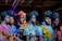 A group of women stands closely together, wearing vibrant, colorful African dresses and headwraps. The outfits feature a variety of patterns, including floral, abstract, and geometric designs. The women have confident expressions and their makeup is elegantly done, accentuating their features. The setting appears to be indoors, possibly under artificial lighting which casts a rich, dramatic effect.