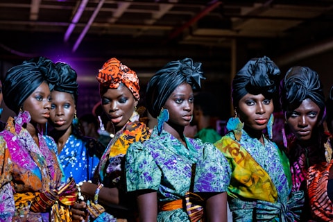 A group of women stands closely together, wearing vibrant, colorful African dresses and headwraps. The outfits feature a variety of patterns, including floral, abstract, and geometric designs. The women have confident expressions and their makeup is elegantly done, accentuating their features. The setting appears to be indoors, possibly under artificial lighting which casts a rich, dramatic effect.