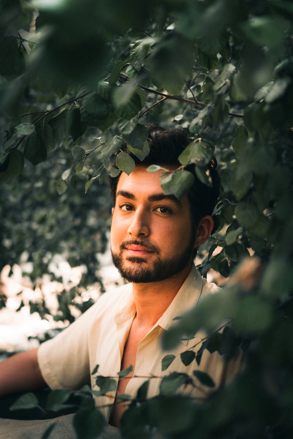 The image shows a young man with short dark hair wearing a white shirt, standing under the leaves of a tree and looking directly at the camera. The background is blurred, drawing focus to the subject's face and upper body.