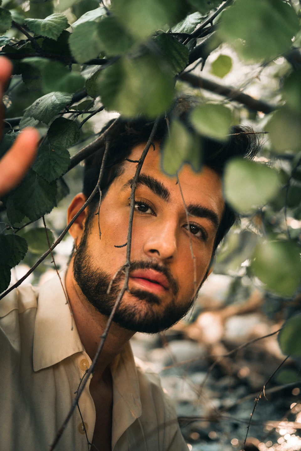 The image shows a young man with a beard and mustache, dressed in a white shirt, peeking out from behind a tree branch. The background is blurred, drawing focus to the subject's face and upper body.