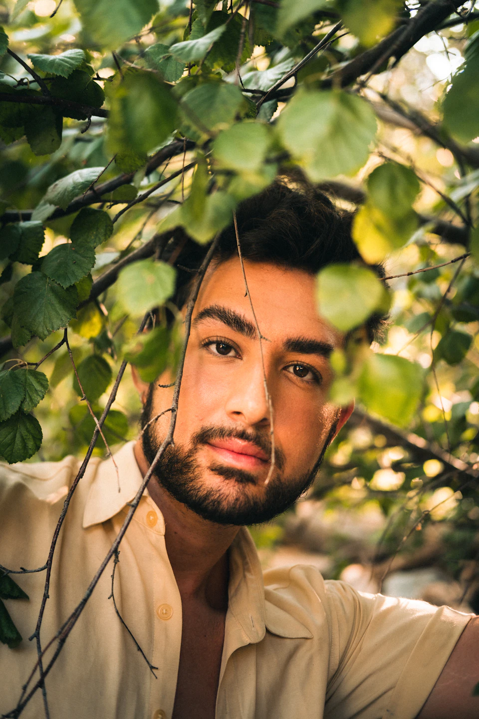 The image shows a young man with a beard and mustache, dressed in a white shirt, peeking out from behind a tree branch. The background is blurred, drawing focus to the subject's face and upper body.