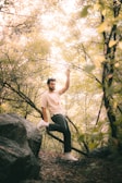 A person is leaning against a large rock surrounded by lush greenery in a forest. Sunlight is filtering through the leaves, creating a soft, dappled lighting effect. The person is wearing casual attire and appears relaxed, immersed in nature.