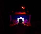 A person is sitting in front of a desk with multiple electronic devices, including a laptop with a glowing screen, and a smartphone. The room is dimly lit with distinctive red and blue lighting, creating a focused and immersive atmosphere. Speakers and other tech gadgets are arranged on the desk.