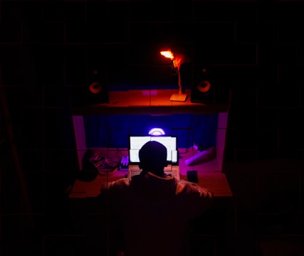A person is sitting in front of a desk with multiple electronic devices, including a laptop with a glowing screen, and a smartphone. The room is dimly lit with distinctive red and blue lighting, creating a focused and immersive atmosphere. Speakers and other tech gadgets are arranged on the desk.