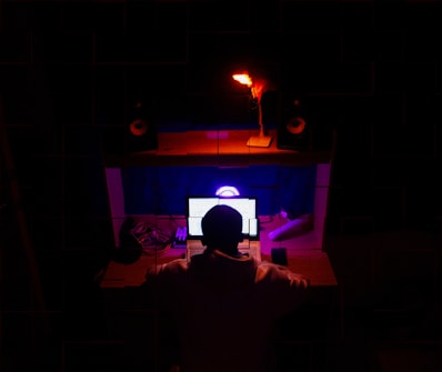 A person is sitting in front of a desk with multiple electronic devices, including a laptop with a glowing screen, and a smartphone. The room is dimly lit with distinctive red and blue lighting, creating a focused and immersive atmosphere. Speakers and other tech gadgets are arranged on the desk.