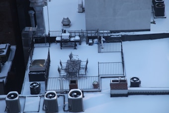 A rooftop is covered with snow, featuring metal railings surrounding patio furniture. The furniture includes a table and several chairs, all accumulating snow. Air conditioning units are dispersed around the rooftop, with snow settling on them as well.