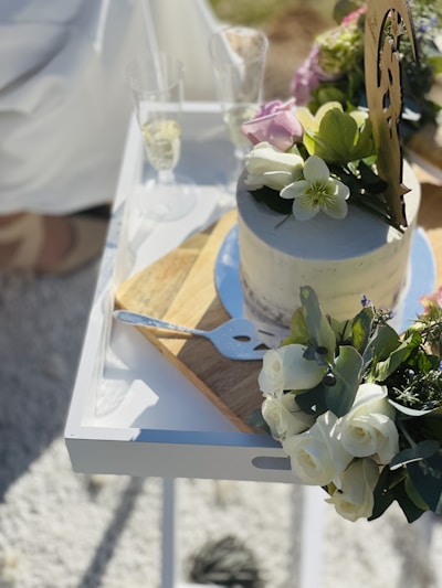A white cake decorated with flowers sits on a wooden board atop a white tray. Next to it are two champagne flutes filled with a light-colored beverage. The cake has flower decorations on the top and nearby there is a cake server. Bright sunlight casts shadows on the textured surface.