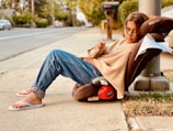 A child in casual clothing leans back comfortably in a Römer car seat placed on the sidewalk, next to a lamppost. The child is wearing a beige sweater, blue jeans, and white flip-flops. A note attached to the car seat reads 'FREE.' The background shows a suburban street with trees, houses, and parked cars, creating a peaceful neighborhood setting.