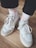 A pair of white sneakers with white laces are worn with artistic white socks featuring red and black designs. The shoes are placed on a wooden floor, and the lower legs are clothed in black pants.
