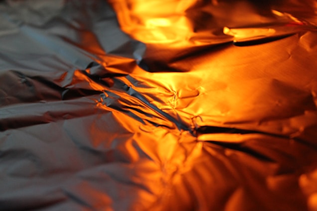 The image features a close-up view of crinkled aluminum foil reflecting warm orange light, creating a textured and abstract pattern.