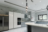 A modern kitchen featuring sleek, white cabinetry and marble-patterned walls. There's a double-door stainless steel refrigerator, an integrated oven, and a microwave. The kitchen island has a smooth, curved design with hanging pendant lights above it. A large window provides natural light, and there's a stainless steel sink with a modern faucet.