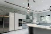 A modern kitchen featuring sleek, white cabinetry and marble-patterned walls. There's a double-door stainless steel refrigerator, an integrated oven, and a microwave. The kitchen island has a smooth, curved design with hanging pendant lights above it. A large window provides natural light, and there's a stainless steel sink with a modern faucet.