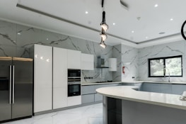 A modern kitchen featuring sleek, white cabinetry and marble-patterned walls. There's a double-door stainless steel refrigerator, an integrated oven, and a microwave. The kitchen island has a smooth, curved design with hanging pendant lights above it. A large window provides natural light, and there's a stainless steel sink with a modern faucet.