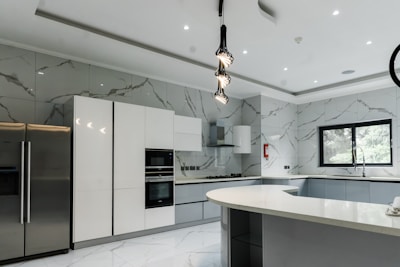 A modern kitchen featuring sleek, white cabinetry and marble-patterned walls. There's a double-door stainless steel refrigerator, an integrated oven, and a microwave. The kitchen island has a smooth, curved design with hanging pendant lights above it. A large window provides natural light, and there's a stainless steel sink with a modern faucet.