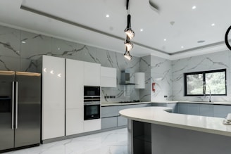A modern kitchen featuring sleek, white cabinetry and marble-patterned walls. There's a double-door stainless steel refrigerator, an integrated oven, and a microwave. The kitchen island has a smooth, curved design with hanging pendant lights above it. A large window provides natural light, and there's a stainless steel sink with a modern faucet.