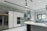 A modern kitchen featuring sleek, white cabinetry and marble-patterned walls. There's a double-door stainless steel refrigerator, an integrated oven, and a microwave. The kitchen island has a smooth, curved design with hanging pendant lights above it. A large window provides natural light, and there's a stainless steel sink with a modern faucet.
