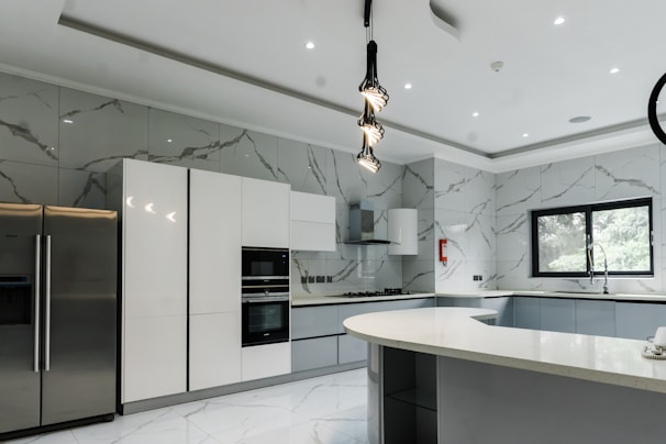 A modern kitchen featuring sleek, white cabinetry and marble-patterned walls. There's a double-door stainless steel refrigerator, an integrated oven, and a microwave. The kitchen island has a smooth, curved design with hanging pendant lights above it. A large window provides natural light, and there's a stainless steel sink with a modern faucet.