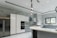 A modern kitchen featuring sleek, white cabinetry and marble-patterned walls. There's a double-door stainless steel refrigerator, an integrated oven, and a microwave. The kitchen island has a smooth, curved design with hanging pendant lights above it. A large window provides natural light, and there's a stainless steel sink with a modern faucet.