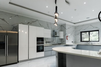 A modern kitchen featuring sleek, white cabinetry and marble-patterned walls. There's a double-door stainless steel refrigerator, an integrated oven, and a microwave. The kitchen island has a smooth, curved design with hanging pendant lights above it. A large window provides natural light, and there's a stainless steel sink with a modern faucet.