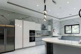 A modern kitchen featuring sleek, white cabinetry and marble-patterned walls. There's a double-door stainless steel refrigerator, an integrated oven, and a microwave. The kitchen island has a smooth, curved design with hanging pendant lights above it. A large window provides natural light, and there's a stainless steel sink with a modern faucet.