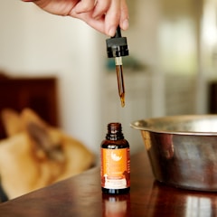 A hand holds a dropper filled with a brown liquid, poised above an open bottle labeled 'slumber sleep aid'. The background features a metallic bowl and blurred objects, giving a warm, cozy ambiance.