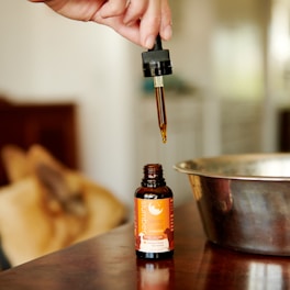 A hand holds a dropper filled with a brown liquid, poised above an open bottle labeled 'slumber sleep aid'. The background features a metallic bowl and blurred objects, giving a warm, cozy ambiance.
