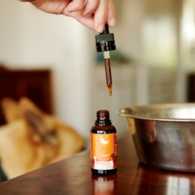 A hand holds a dropper filled with a brown liquid, poised above an open bottle labeled 'slumber sleep aid'. The background features a metallic bowl and blurred objects, giving a warm, cozy ambiance.