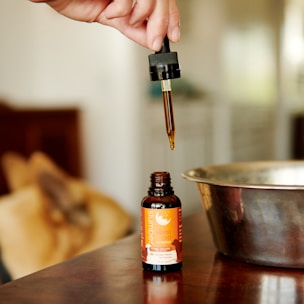 A hand holds a dropper filled with a brown liquid, poised above an open bottle labeled 'slumber sleep aid'. The background features a metallic bowl and blurred objects, giving a warm, cozy ambiance.