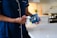 A person wearing navy blue pajamas stands in a modern kitchen, holding a packet of sleep aid gummies. The kitchen features a white marble countertop and a patterned backsplash. A stove is visible in the background along with a small plant.