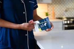 A person wearing navy blue pajamas stands in a modern kitchen, holding a packet of sleep aid gummies. The kitchen features a white marble countertop and a patterned backsplash. A stove is visible in the background along with a small plant.