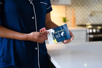 A person wearing navy blue pajamas stands in a modern kitchen, holding a packet of sleep aid gummies. The kitchen features a white marble countertop and a patterned backsplash. A stove is visible in the background along with a small plant.