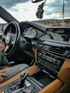 The interior of a car with a focus on the dashboard and driver's area. The dashboard features a digital display and various control buttons. A driver's hand rests on the steering wheel. The interior has brown leather upholstery and a sleek, modern design. A scenic view of a landscape with buildings and a cloudy sky is visible through the window.