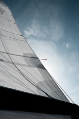 A close-up view of a sailing boat's sail against a partly cloudy sky. The sail is stretched taut, with visible ropes and stitching patterns. The sun filters through the fabric, giving it a translucent quality.