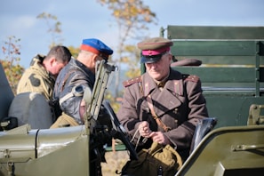 A group of individuals dressed in military-style attire are positioned around and inside an old-fashioned military vehicle. The person in the foreground wears a brown uniform with a red and brown hat, suggesting a vintage military context. Another individual is wearing a cap with aviator goggles pushed up to the forehead. The vehicle is painted in army green, and there are trees in the background indicating an outdoor setting.