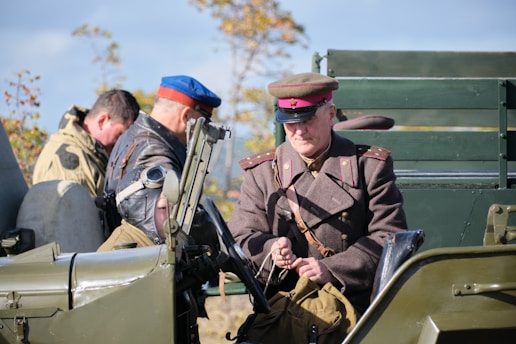 A group of individuals dressed in military-style attire are positioned around and inside an old-fashioned military vehicle. The person in the foreground wears a brown uniform with a red and brown hat, suggesting a vintage military context. Another individual is wearing a cap with aviator goggles pushed up to the forehead. The vehicle is painted in army green, and there are trees in the background indicating an outdoor setting.