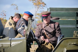 A group of individuals dressed in military-style attire are positioned around and inside an old-fashioned military vehicle. The person in the foreground wears a brown uniform with a red and brown hat, suggesting a vintage military context. Another individual is wearing a cap with aviator goggles pushed up to the forehead. The vehicle is painted in army green, and there are trees in the background indicating an outdoor setting.