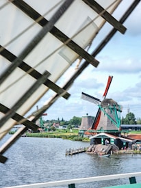 A scenic view featuring traditional Dutch windmills near a calm water body under a partly cloudy sky. One windmill is prominently visible with a thatched roof and green trim, with another in the distance. The landscape is lush with green vegetation and small houses.