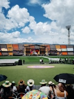 A sports stadium with a large stage set up on the field, hosting an outdoor event. Spectators wearing colorful hats sit in the foreground. The seating areas display vibrant colors, and a clear blue sky with scattered clouds is visible. Large screens display graphics related to the event.