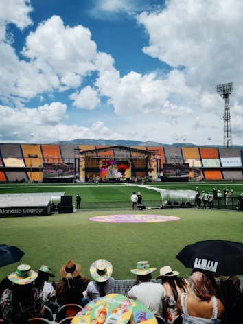 A sports stadium with a large stage set up on the field, hosting an outdoor event. Spectators wearing colorful hats sit in the foreground. The seating areas display vibrant colors, and a clear blue sky with scattered clouds is visible. Large screens display graphics related to the event.